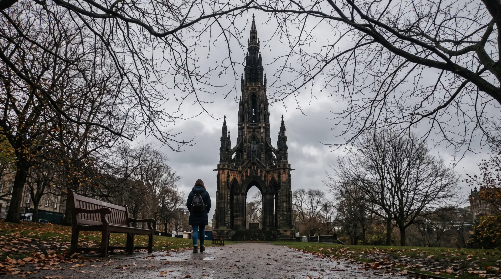 The blackened Gothic spire of the Scott Monument rising over East Princes Street Gardens under flat grey cloud, seen from the gravel path below in Edinburgh